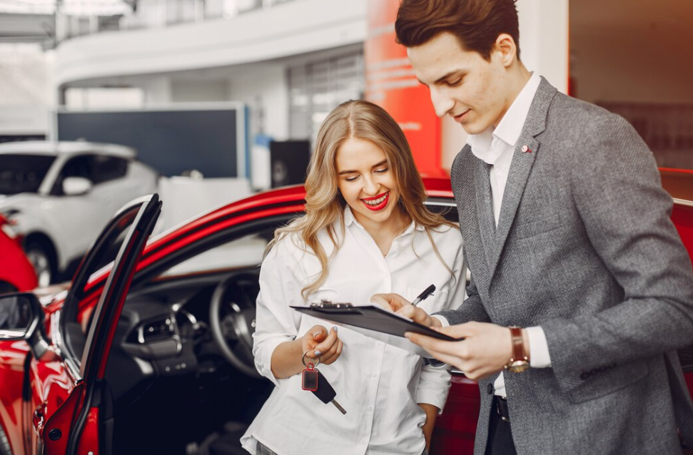 A woman holding car keys and a man holding clipboard and stand beside a red vehicle indoors.